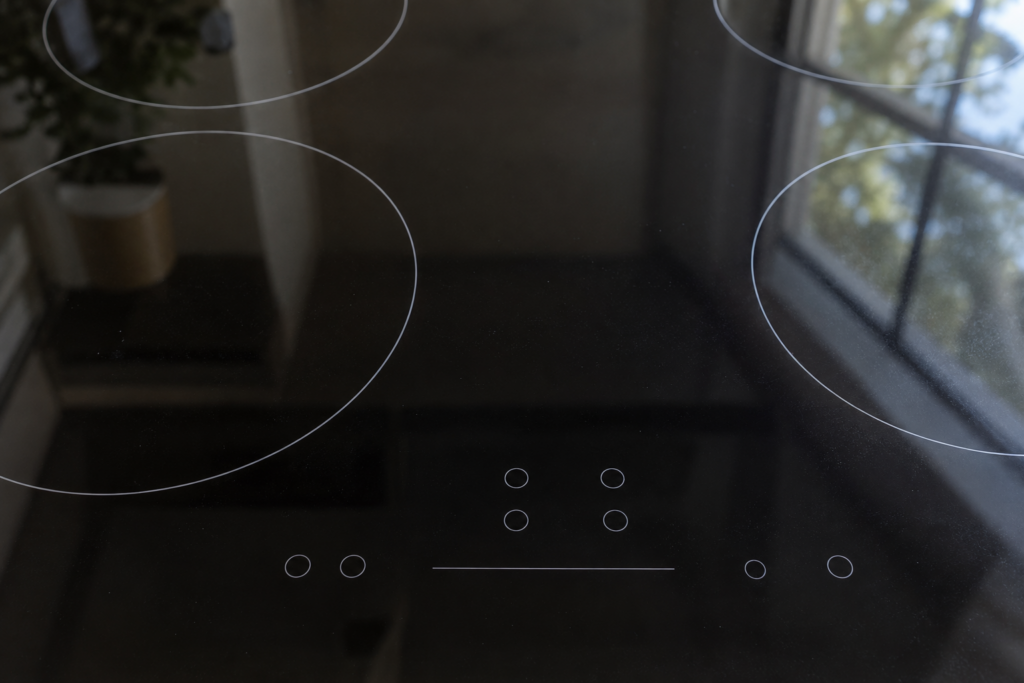 Macro of an induction cooktop's touch control glass surface from above, showing a faint reflection of a kitchen window.