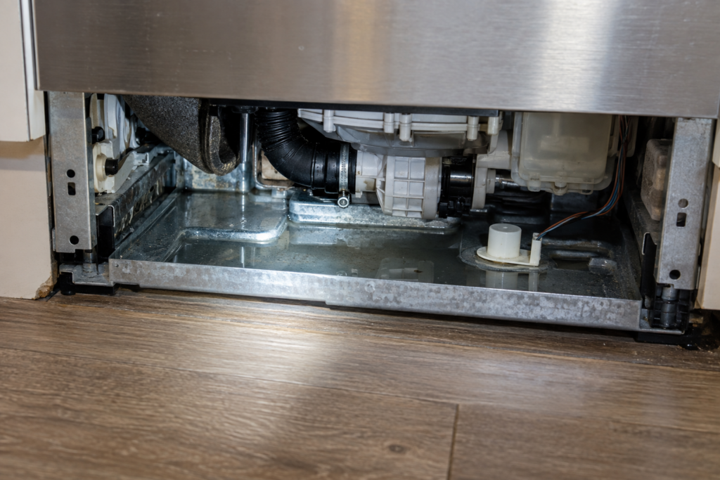 Close-up under a dishwasher with its lower kickplate removed, showing a metal base pan with a small pool of water and a float sensor beside it.