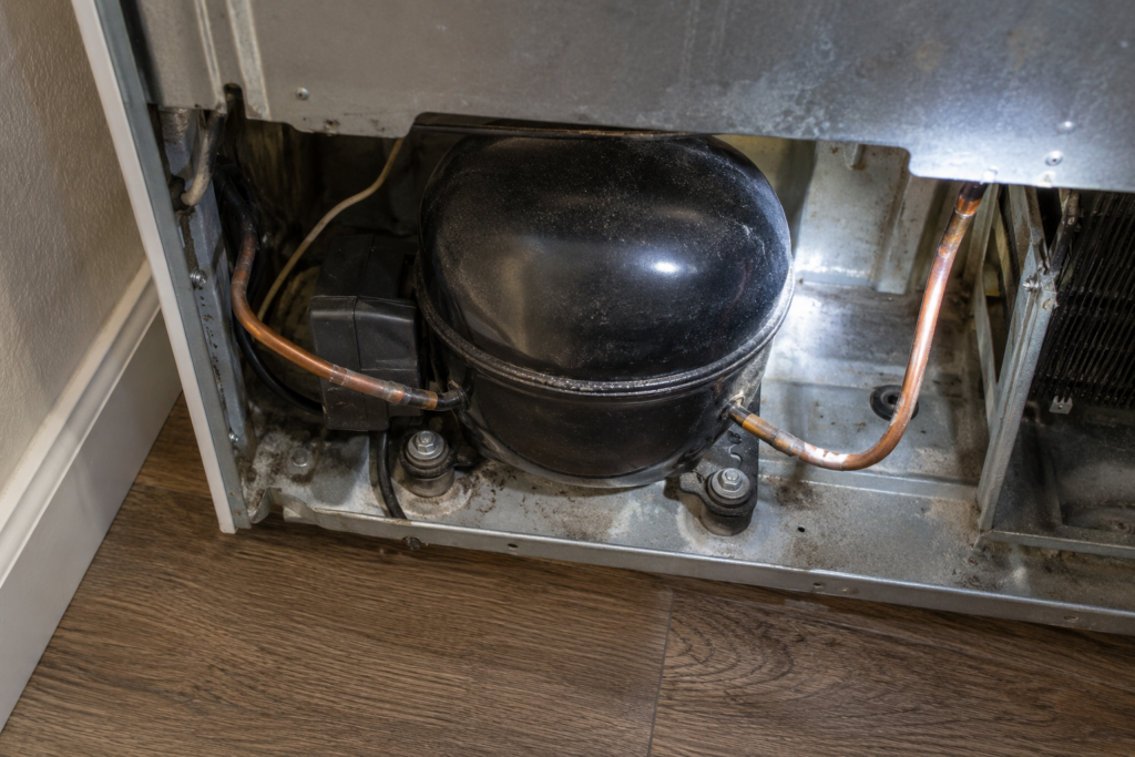 A black compressor dome with two copper tubes and a small relay box seen from above in the bottom rear compartment of a refrigerator pulled away from the wall, a flashlight beam from the right.