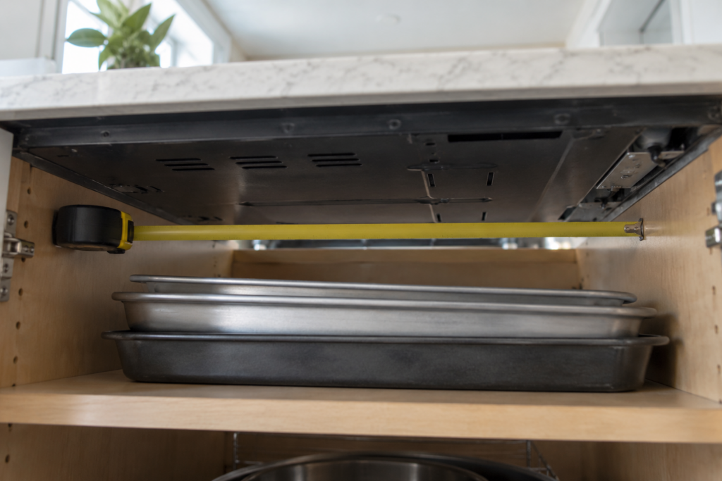 Looking up into the open base cabinet beneath an induction cooktop, a tape measure laid across the gap below the cooktop underside and a few stored baking trays sitting too close to it, daylight from the kitchen.